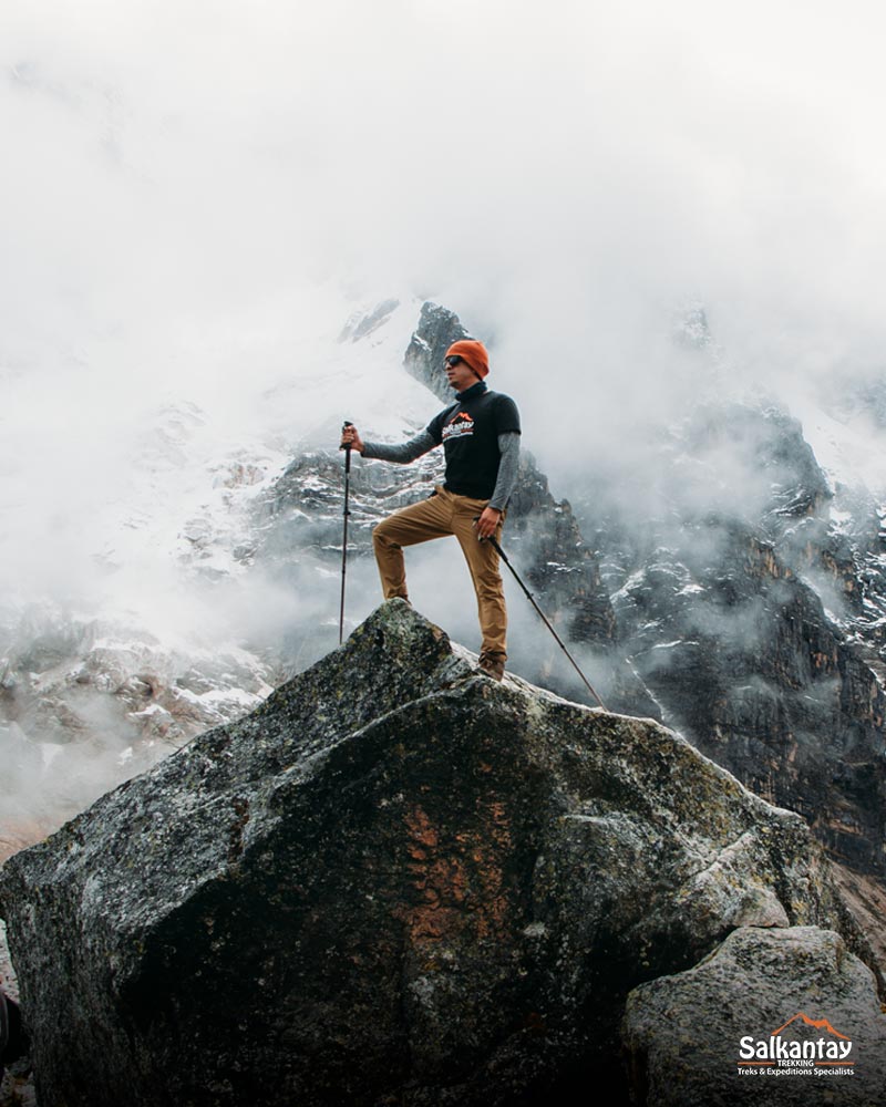 Turista en lo alto de una roca gigante con bastones de senderismo en el abra de Salkantay