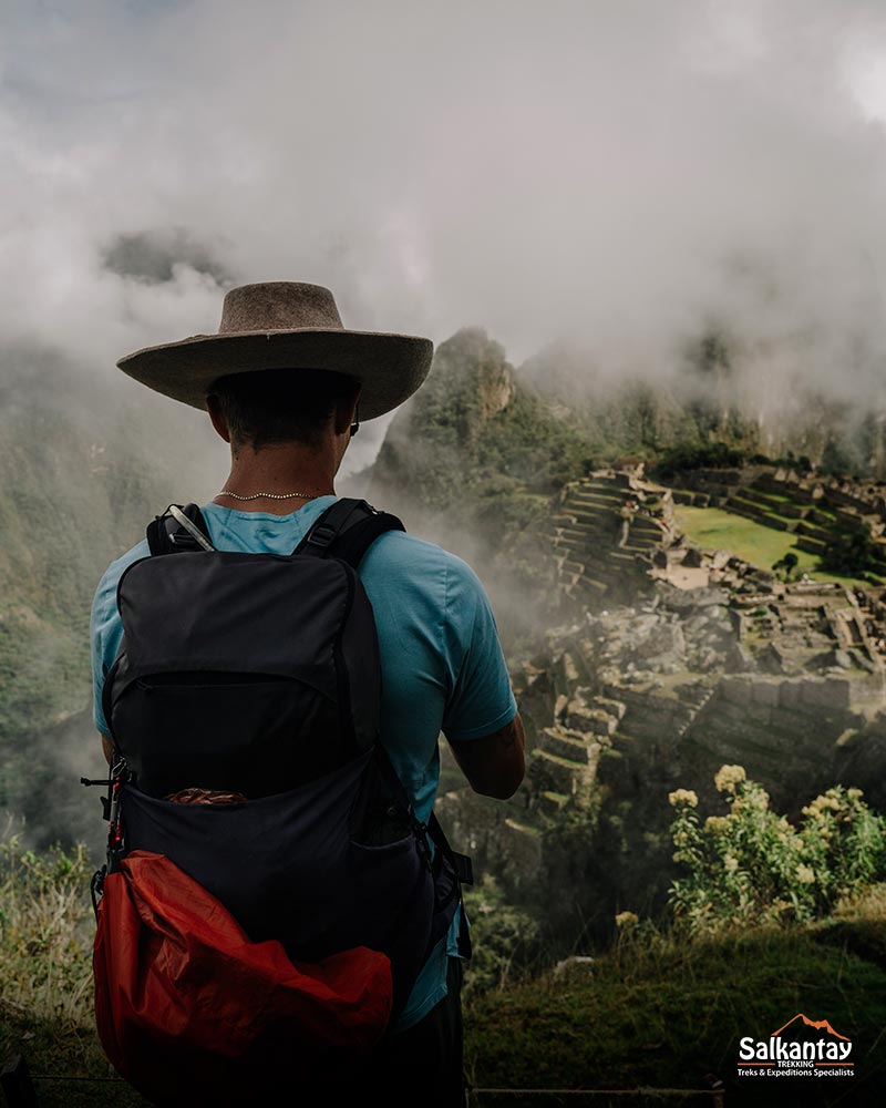 Turista masculino con sombrero andino admirando la ciudad inca de Machu Picchu llena de nubes.