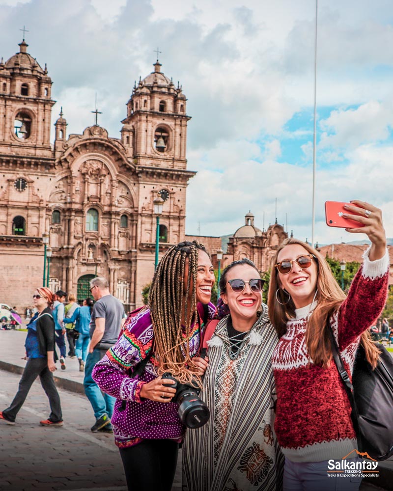 Tres turistas en la plaza principal de Cusco sonriendo y tomándose un selfie con sus amigas.