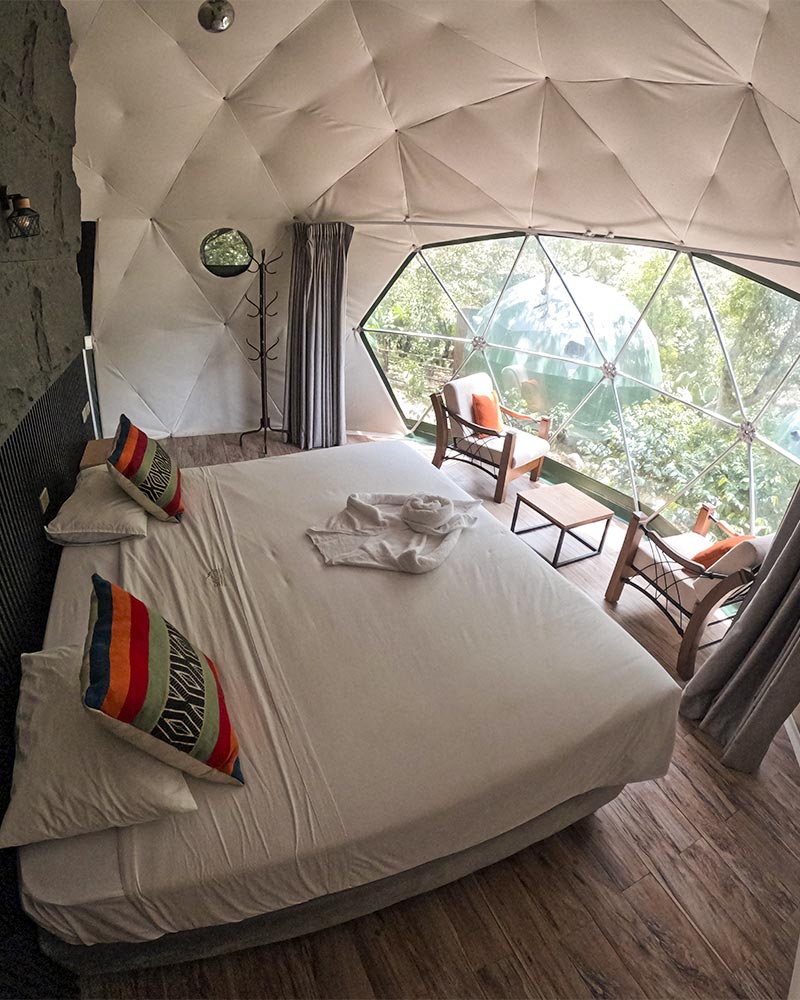 Campamento de lujo, glamping Super Jungle Domes por dentro, cama espaciosa, sillas, mesa de centro y vista panoramica de la naturaleza