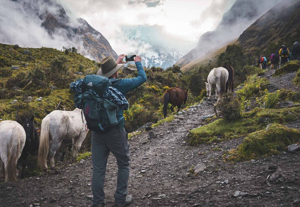Turista tomando una foto con su celular en la ruta Salkantay, rodeado de vegetación, neblina y caballos