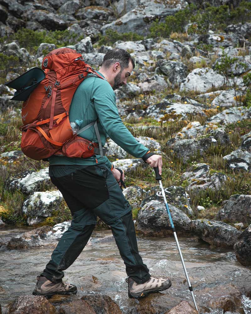 Senderista cruzando un arroyo en la caminata Salkantay, con palos de trekking y una mochila que se ajusta bien a su contextura