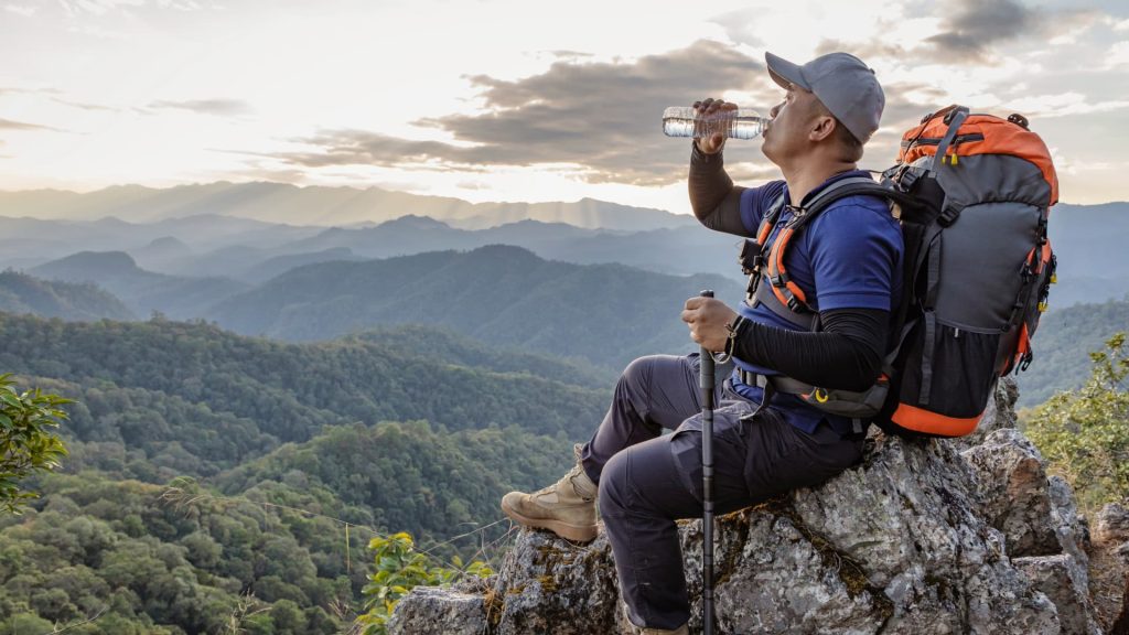 Un excursionista bebe agua de una botella mientras está de pie con dos amigos en un sendero rocoso de montaña con picos nevados.