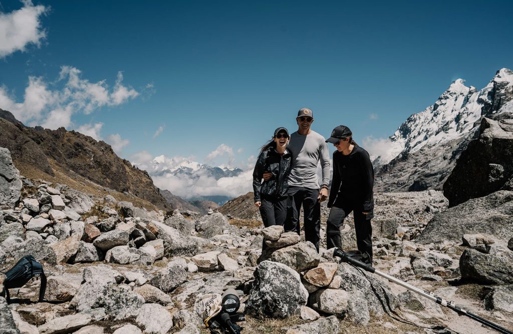 Tres excursionistas sonriendo en un sendero rocoso de montaña con picos nevados al fondo.