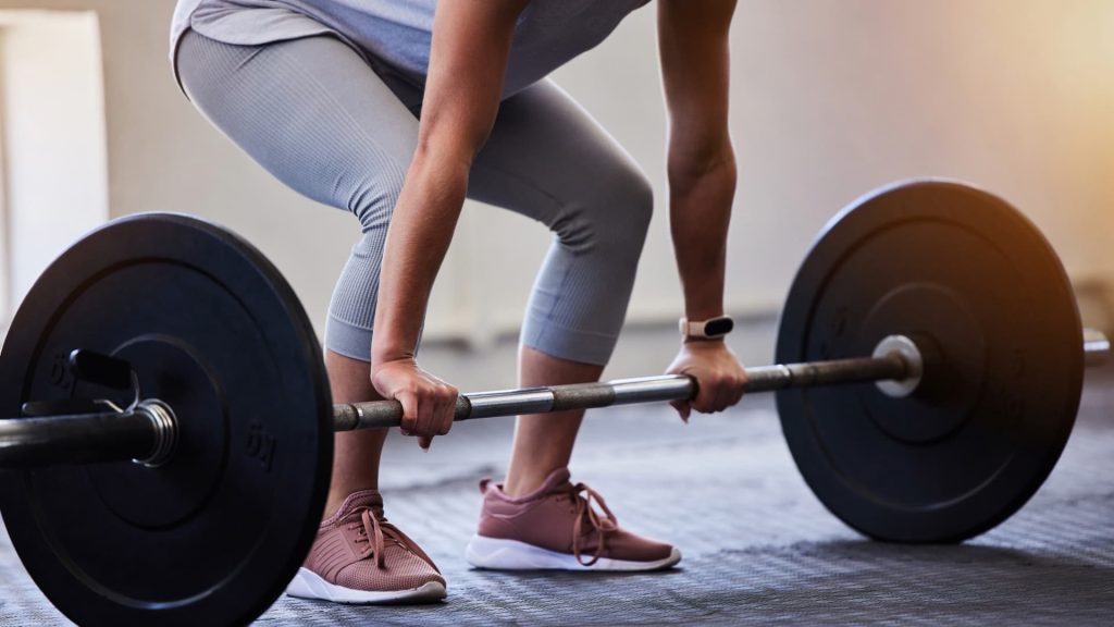 Primer plano de una persona sujetando una barra cargada, preparándose para realizar un ejercicio de peso muerto en un gimnasio.