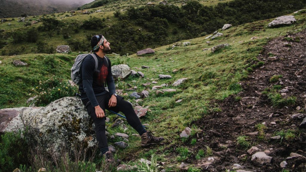 Una persona cansada, con una mochila a la espalda, descansando sobre una roca durante una exigente ruta de montaña a gran altitud.