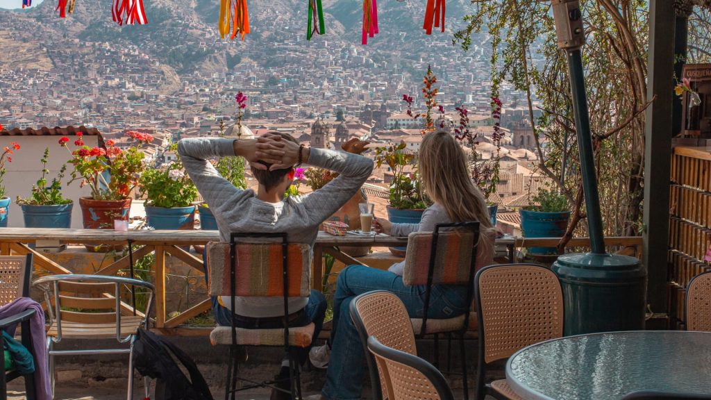 Una vista panorámica de la animada Plaza de Armas de Cuzco, Perú, enmarcada por las ventanas de madera de la terraza del balcón de un restaurante situado en la segunda planta, en la que se aprecia la arquitectura colonial y las montañas bajo un cielo azul.
