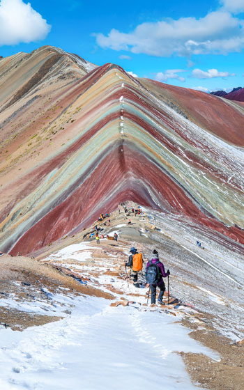 Dos turistas caminando frente a la Montaña de Siete Colores (Vinicunca) en Perú. Paisaje vibrante de los Andes con colinas multicolores.