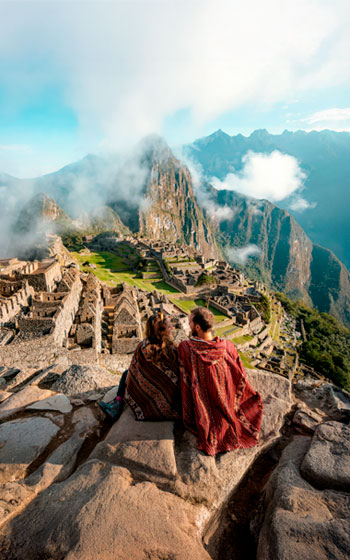 Pareja de turistas con ponchos andinos disfrutando la vista de la ciudadela inca de Machu Picchu, rodeada de montañas y un hermoso paisaje andino.