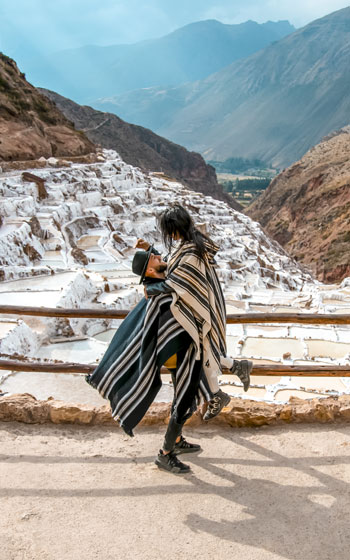 Pareja de turistas con ponchos andinos en las Salineras de Maras, admirando las terrazas de sal tradicionales en medio de los paisajes montañosos del Valle Sagrado.