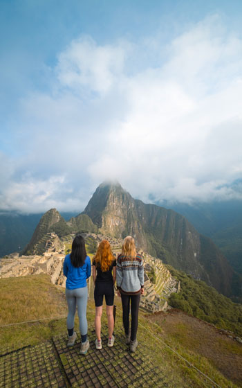 Tres turistas observan la majestuosa ciudadela inca de Machu Picchu, rodeada de montañas verdes y un paisaje andino impresionante.