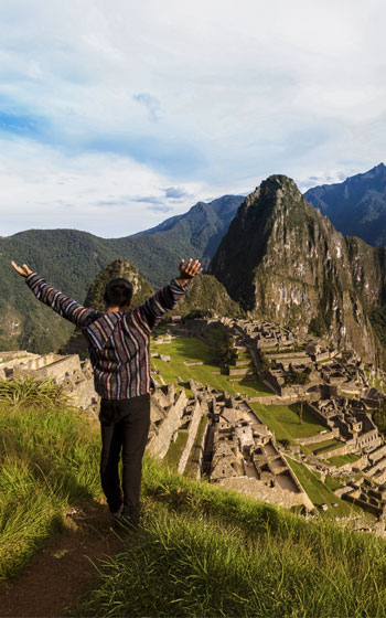 Turista levantando los brazos mientras contempla la impresionante ciudadela inca de Machu Picchu, rodeada de montañas y un paisaje andino espectacular.