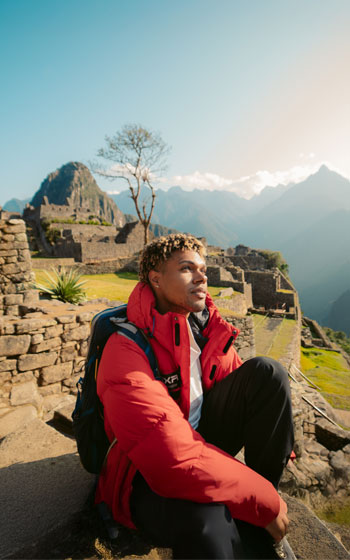 Turista con chaqueta roja posando en primer plano frente a las ruinas de la ciudadela inca de Machu Picchu.