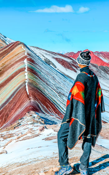 Turista con poncho andino contemplando la Montaña de Colores, rodeada de vibrantes tonalidades y paisajes impresionantes de los Andes.