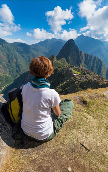 Turista sentado contemplando la vista panorámica de la ciudadela inca de Machu Picchu, rodeada de montañas y un paisaje andino impresionante.