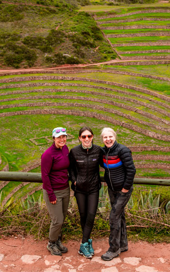 Tres turistas posan con el impresionante laboratorio agrícola de Moray al fondo, destacando sus terrazas circulares y el paisaje andino del Valle Sagrado.