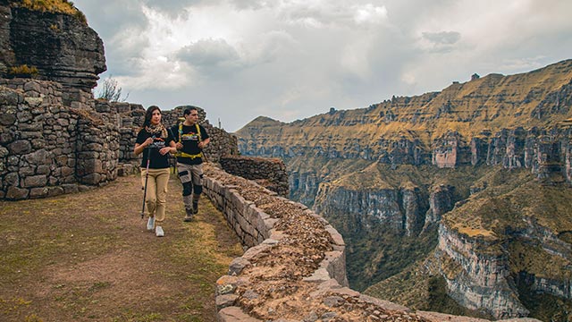 Pareja de turistas sentados con el imponente fondo del complejo arqueológico de Waqrapukara, rodeado de formaciones rocosas y paisajes andinos.