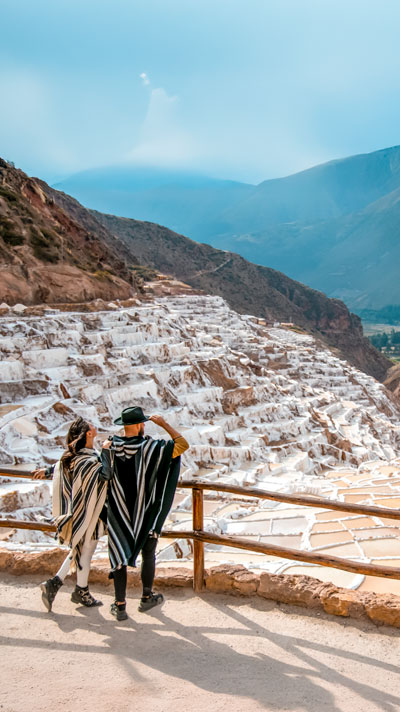 Pareja de turistas con ponchos andinos en las Salineras de Maras, enclavadas en las montañas de los Andes sobre el Valle Sagrado de los Incas, admirando sus antiguas pozas de sal.