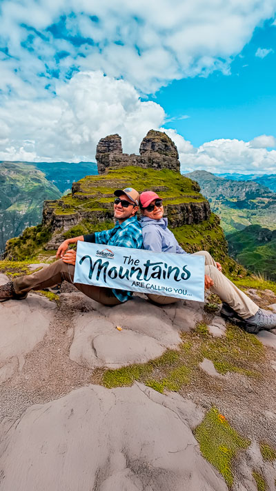 Pareja de turistas sentados con el imponente fondo del complejo arqueológico de Waqrapukara, rodeado de formaciones rocosas y paisajes andinos.