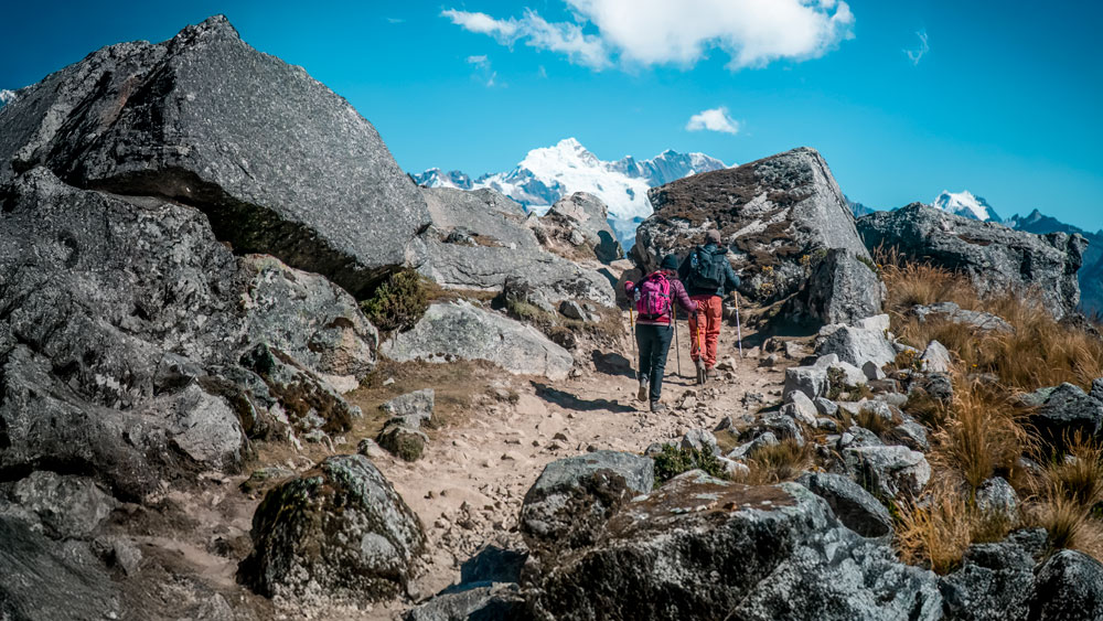 Dos senderistas con mochilas y bastones subiendo por un sendero rocoso hacia montañas nevadas en el Salkantay Trek