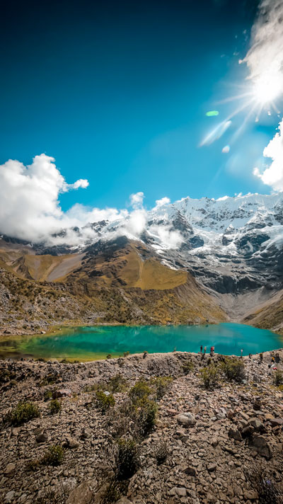 Vista panorámica de turistas admirando la asombrosa Laguna Humantay bajo la imponente Montaña Humantay, rodeada de paisajes andinos.