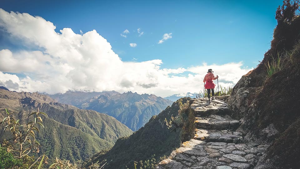 Senderista con bastones subiendo escalones de piedra del Camino Inca con un valle y el cielo azul de fondo