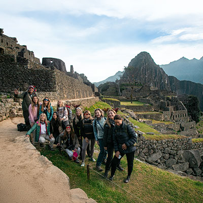 Grupo de turistas sonrientes posando para una foto grupal en el sector de la Casa del Guardián con la ciudadela de Machu Picchu de fondo.