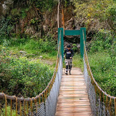 Turista cruzando un puente colgante de madera y metal sobre el caudaloso río Urubamba en el trayecto hacia Aguas Calientes, Cusco.