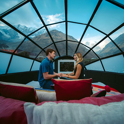Pareja de viajeros descansando dentro de un domo de vidrio (Sky Camp) con vista directa al majestuoso nevado Salkantay y la montaña Humantay en Cusco.