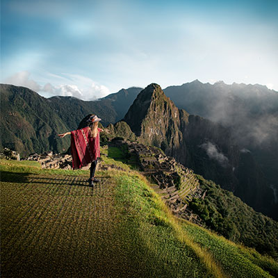 Turista posando con un poncho andino de colores vibrantes frente a la ciudadela inca de Machu Picchu y la montaña Huayna Picchu en Cusco, Perú.