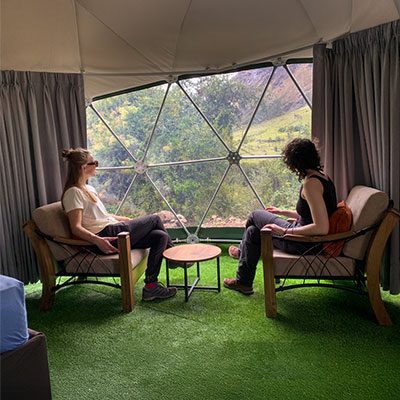 Una pareja de turistas sentados y relajándose dentro de un cómodo domo geodésico en el campamento Salkantay Mountain Domes, con una vista panorámica de las montañas visible a través del ventanal.